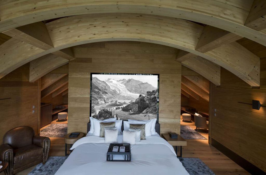 Modernes Schlafzimmer mit gewölbter Holzdecke, einem Bett mit weißer Bettwäsche, einem großen Bergbild an der Wand und einem Ledersessel.