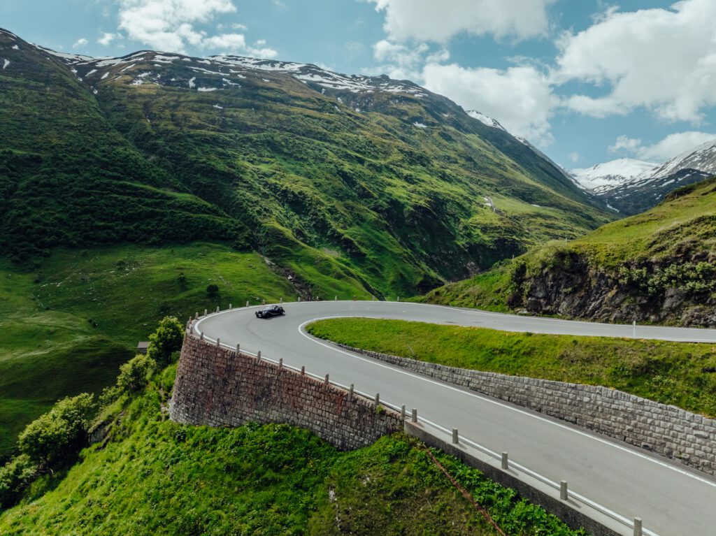 Ein Auto fährt auf einer kurvenreichen Straße durch üppige, grüne Berge unter einem teilweise bewölkten Himmel.