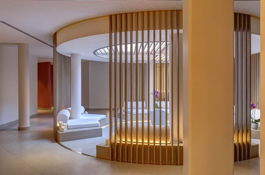 Moderner, runder Sitzbereich mit weißen Polsterbänken, umgeben von vertikalen Holzlatten. Sanfte Beleuchtung unterstreicht die ruhige Atmosphäre. Lila Orchideen stehen in der Mitte.