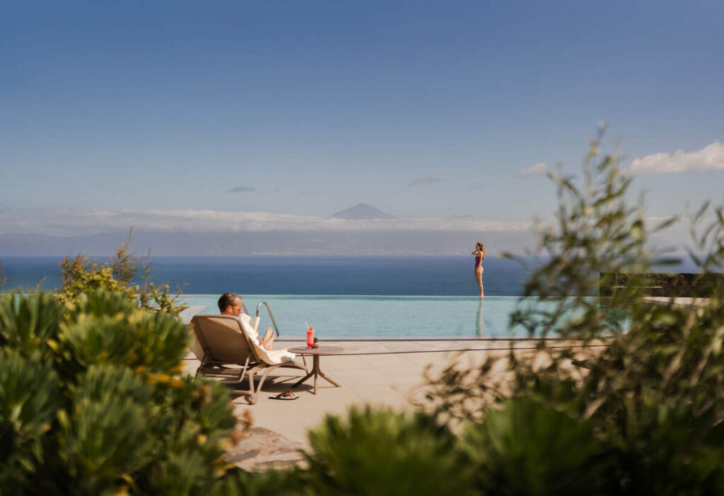Eine Person entspannt sich auf einem Liegestuhl neben einem Infinity-Pool, während eine andere am Beckenrand steht; im Hintergrund sind in der Ferne Berge und ein blauer Himmel zu sehen.