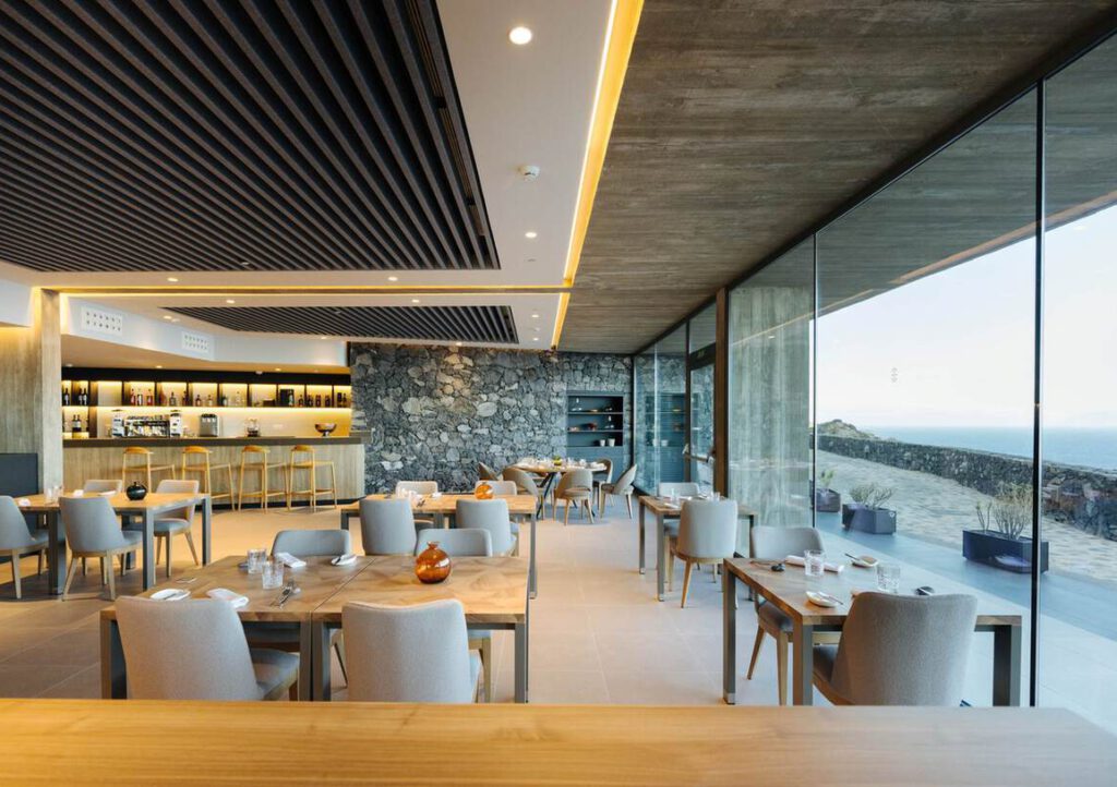 Modernes Restaurant-Interieur mit Holztischen, gepolsterten Stühlen, einer Akzentwand aus Stein und großen, vom Boden bis zur Decke reichenden Fenstern mit Blick auf eine Außenterrasse und eine malerische Landschaft.