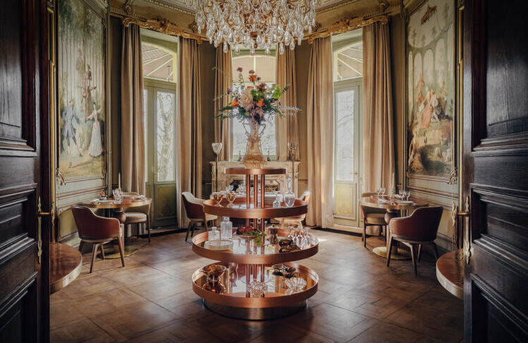 Eleganter Speisesaal mit kunstvollem Kronleuchter, klassischen Wandgemälden, großen Fenstern, runden Tischen und einem zentralen Etagerenständer mit Glaswaren und Blumen.