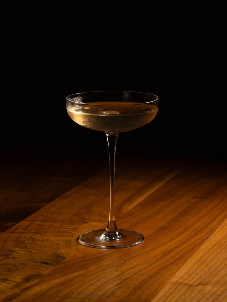 Ein mit einem klaren, hellen Getränk gefülltes Coupe-Glas steht auf einer Holzfläche vor einem dunklen Hintergrund.