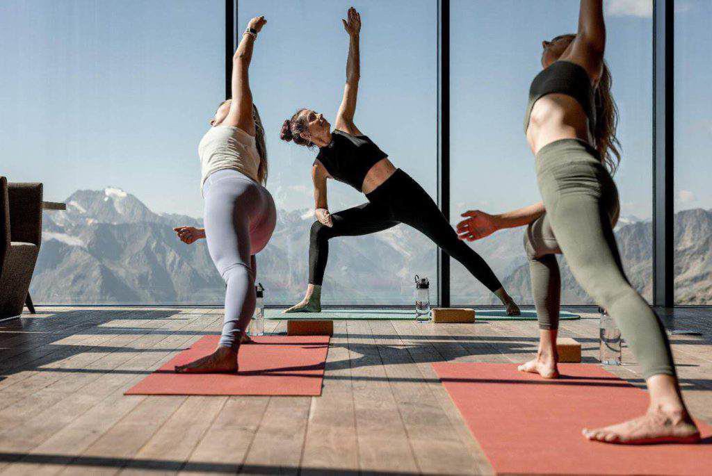 Drei Personen praktizieren Yoga auf Matten in einem hellen Raum mit großen Fenstern und Blick auf eine malerische Berglandschaft.