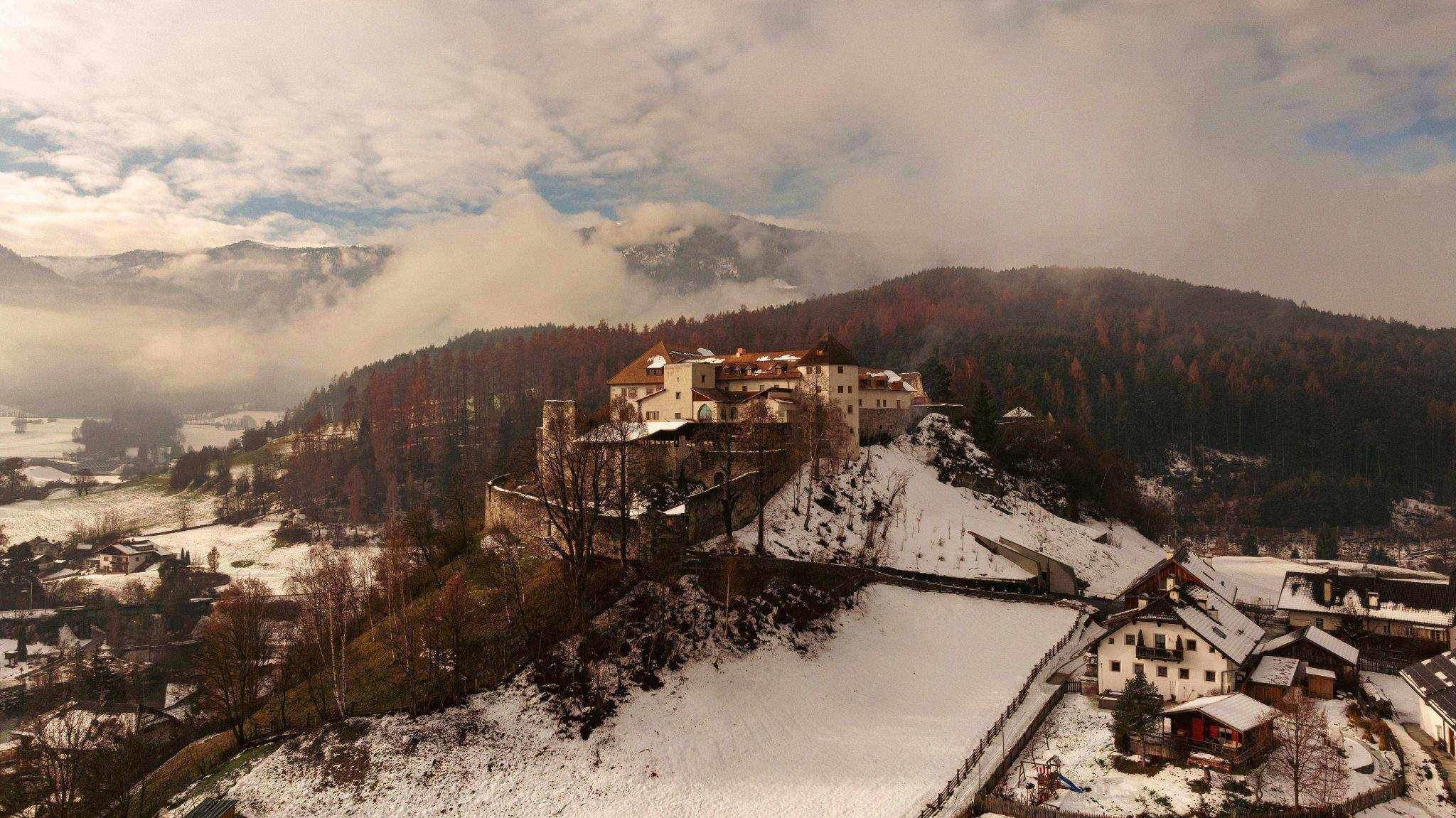 Eine Luftaufnahme eines großen, befestigten Schlosses auf einem Hügel, das von tiefem, schneebedecktem Wald und einem kleinen Dorf am Fuße des Hügels umgeben ist, unter einem teilweise bewölkten Himmel.