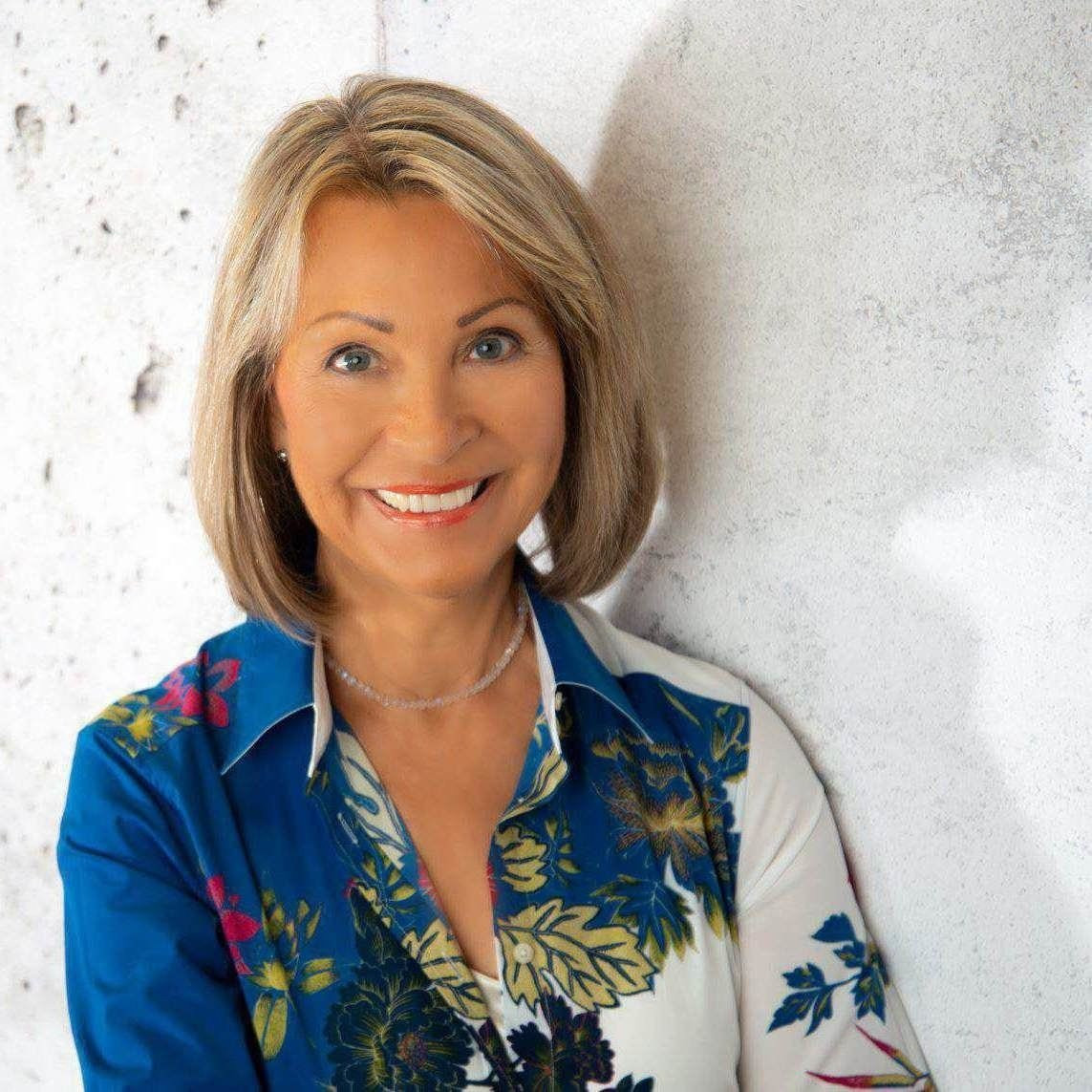 Eine Frau mit schulterlangem blondem Haar und geblümter Bluse steht lächelnd vor einer hellen, strukturierten Wand.