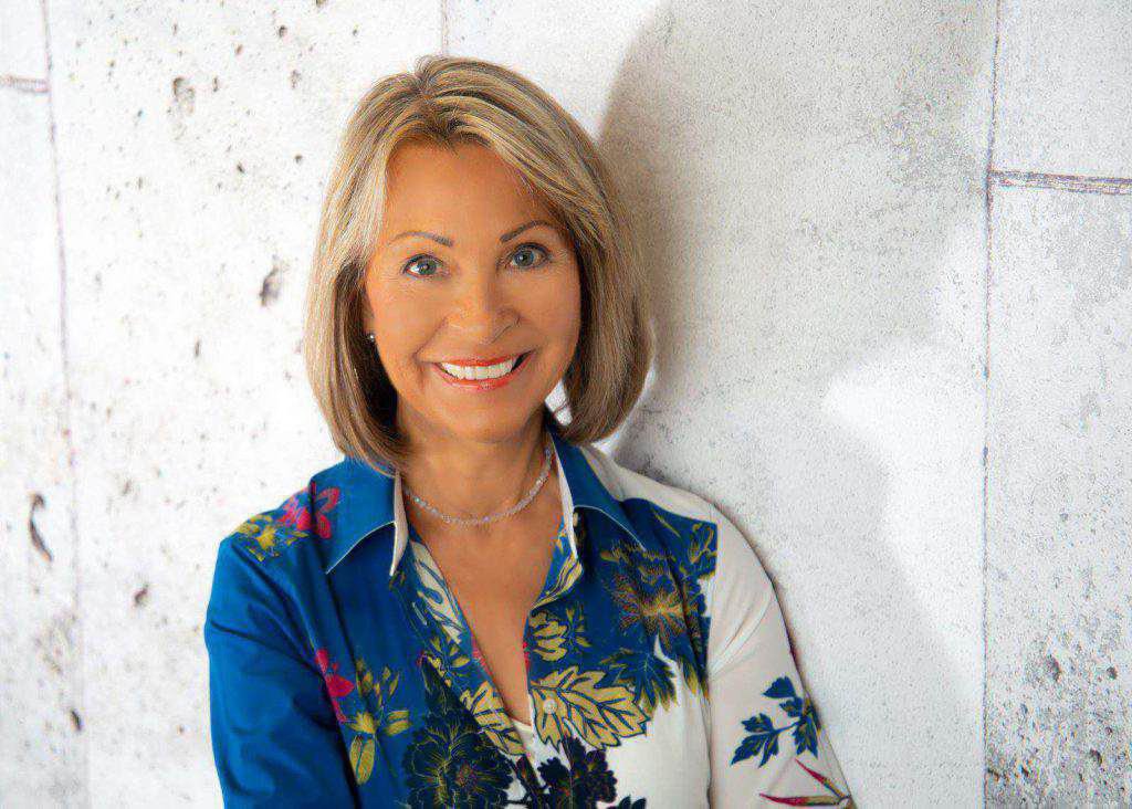 Heike Neuenburg, mit schulterlangen blonden Haaren und blau-weiß geblümter Bluse, steht mit einem warmen Lächeln vor einer hellen, strukturierten Wand.