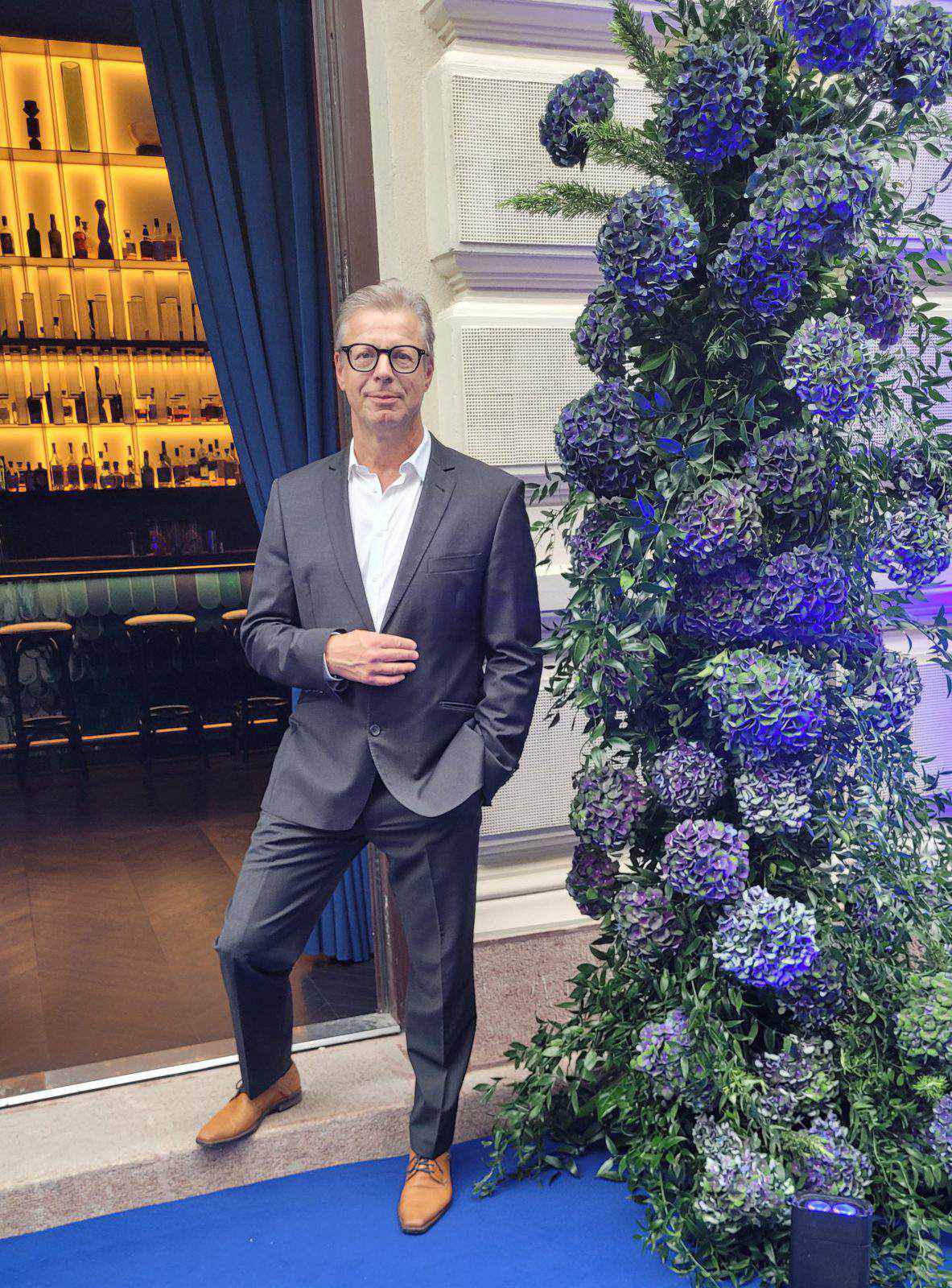 Udo Schwickal, im grauen Anzug und mit braunen Schuhen, steht auf einem blauen Teppich neben einem großen Blumenarrangement, im Hintergrund ist eine Bar zu sehen - perfekt für ein Autorenprofil-Foto.