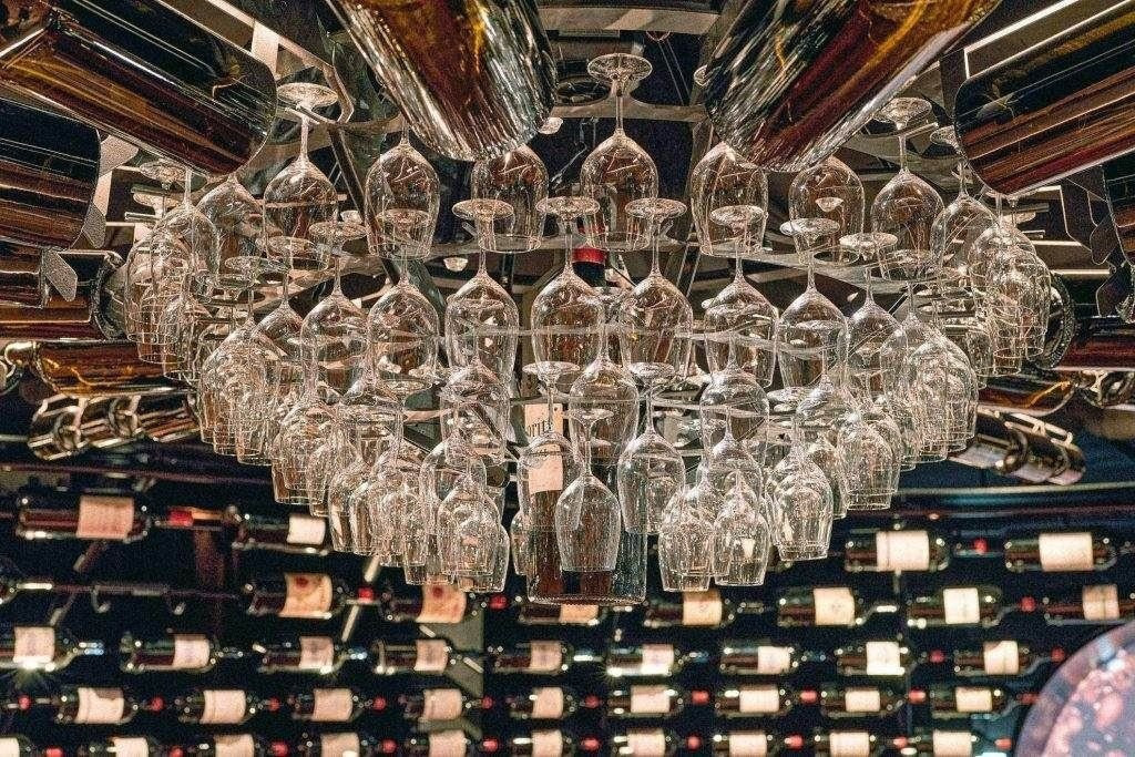 Reihen von Weingläsern hängen kopfüber von einem Regal, während im Hintergrund Regale mit Weinflaschen an der Wand angebracht sind - das perfekte LuxSpots-Ambiente für Weinliebhaber.