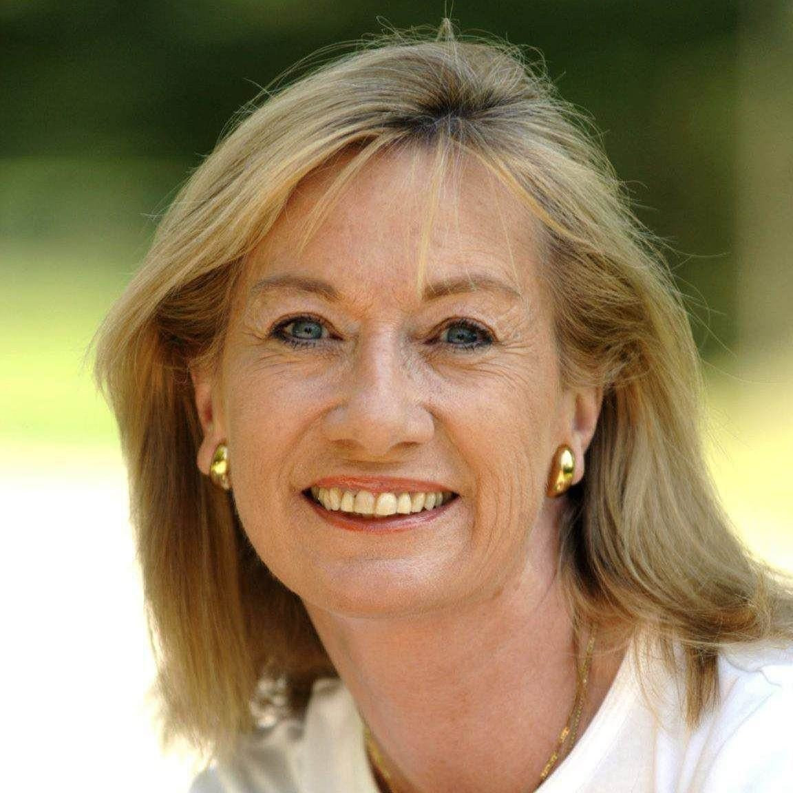 Eine Frau mittleren Alters mit blondem Haar und blauen Augen lächelt im Freien, trägt ein weißes Oberteil und goldene Ohrringe. Der Hintergrund ist unscharfes Grün.