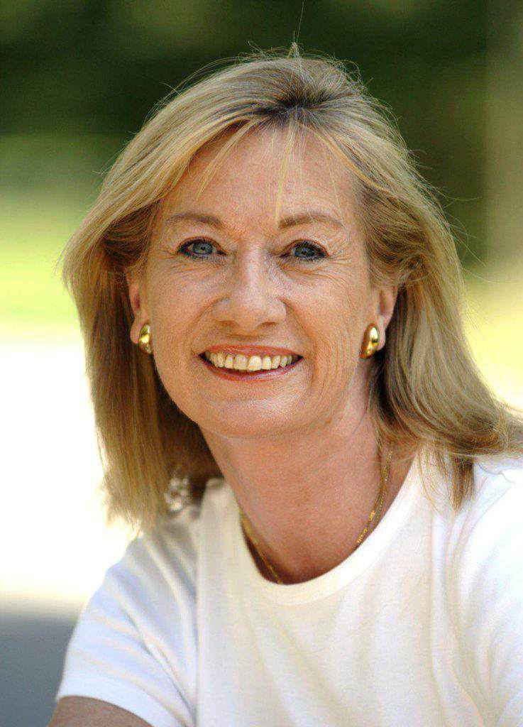 Autorin Claudia Bette-Wenngatz lächelt mit blondem Haar, weißem T-Shirt und goldenen Ohrringen im Freien in die Kamera.