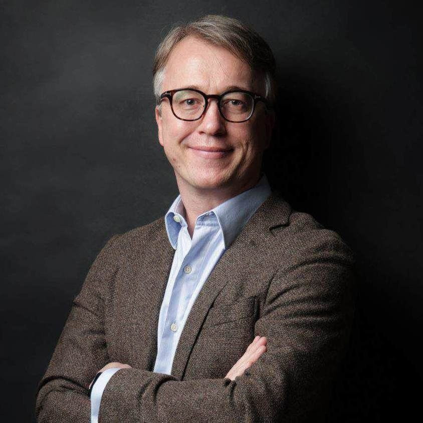 Ein Mann mittleren Alters mit Brille und grauem Haar, der einen braunen Blazer und ein hellblaues Hemd trägt, steht mit verschränkten Armen vor einem dunklen Hintergrund.