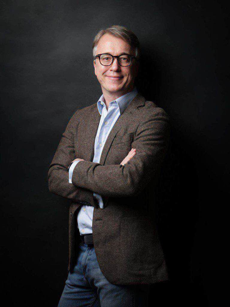 Autor und LuxSpots-Herausgeber, Sascha Ihns, steht mit verschränkten Armen in braunem Blazer, hellblauem Hemd und Jeans vor einem schlichten dunklen Hintergrund.