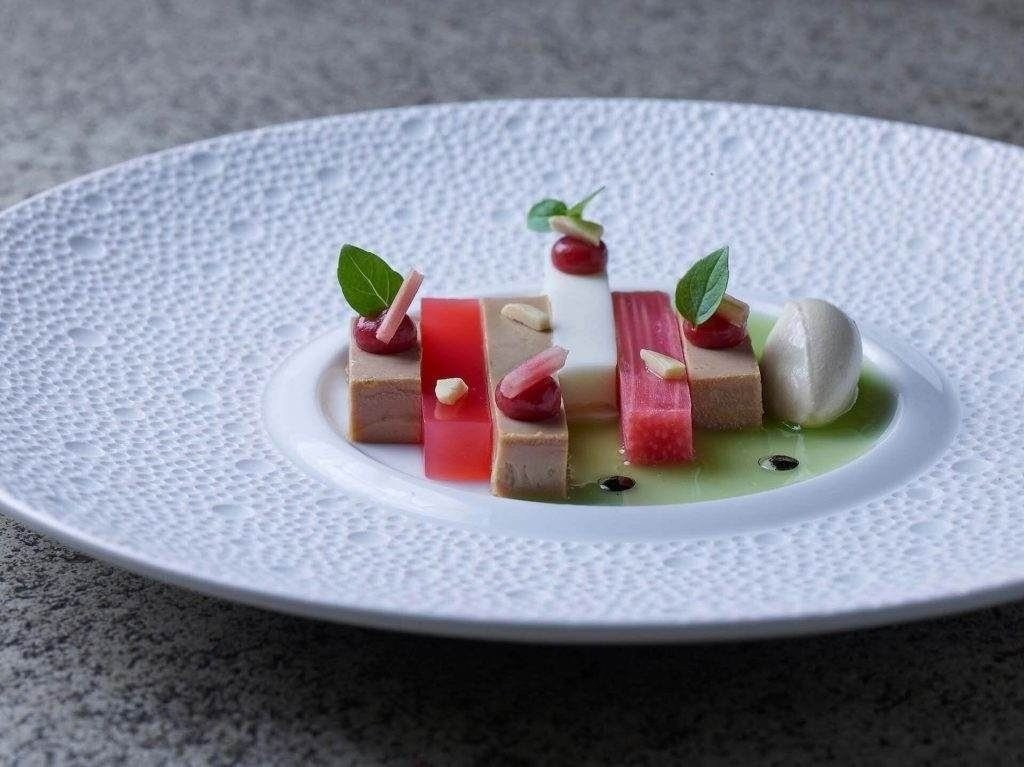 Ein Gourmetteller mit rechteckigen Schichten aus Terrine, rotem Gelee und Panna Cotta, garniert mit Soßentupfen, Mikrogrün, Mandelsplittern und einem Quäntchen Sorbet - perfekt, um es bei LuxSpots zu entdecken.