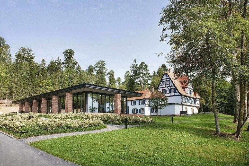 Ein modernes Gebäude aus Glas und Stein steht neben einem traditionellen Fachwerkhaus mit rotem Dach, umgeben von grünen Rasenflächen und Bäumen - ein architektonischer Kontrast, der den einzigartigen Charme von LuxSpots unterstreicht.